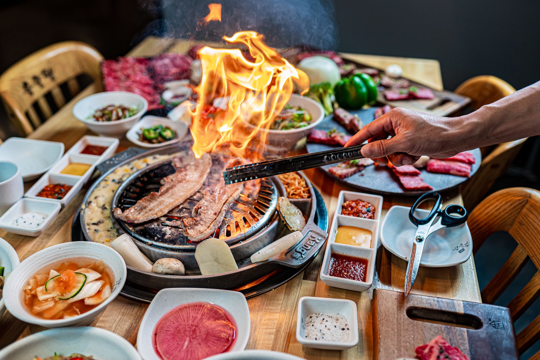Flames rising from a Korean barbecue grill loaded with meat and side dishes on a wooden table